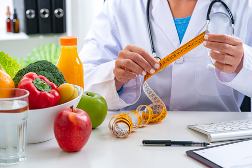 Right nutrition and diet concept. Nutritionist sitting at her desk holding a red apple explaining the benefits of eating fruits and vegetables. An orange juice bottle, glass of water, tape measure are on the doctor's desk and complete the composition. Copy space available for text and/or logo. High resolution 42Mp studio digital capture taken with Sony A7rII and Sony FE 90mm f2.8 macro G OSS lens