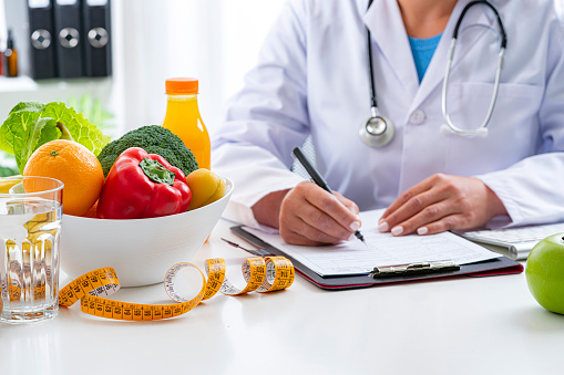 Right nutrition and diet concept. Nutritionist sitting at her desk writing prescription and explaining the benefits of eating fruits and vegetables. An orange juice bottle, glass of water, tape measure are on the doctor's desk and complete the composition. Copy space available for text and/or logo. High resolution 42Mp studio digital capture taken with Sony A7rII and Sony FE 90mm f2.8 macro G OSS lens