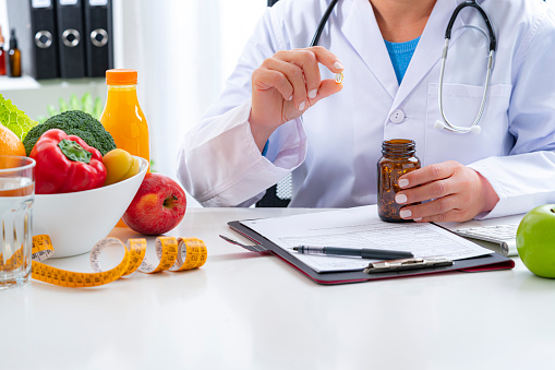 Right nutrition and diet concept. Nutritionist sitting at her desk holding a nutritional supplement capsule and bottle and explaining the benefits of eating fruits and vegetables. An orange juice bottle, glass of water, tape measure are on the doctor's desk and complete the composition. Copy space available for text and/or logo. High resolution 42Mp studio digital capture taken with Sony A7rII and Sony FE 90mm f2.8 macro G OSS lens