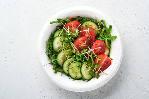 Fresh salad with tomato, cucumber, vegetables, microgreen radishes in white plate on grey stone background. View from above. Concept vegan and healthy eating.
