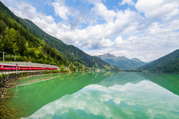 A red train of the Rhaetian Railway passing on the shore of Lago di Poschiavo, a natural lake in the Val di Poschiavo in the Swiss canton of Graubunden. Bernina range reflected in the water.