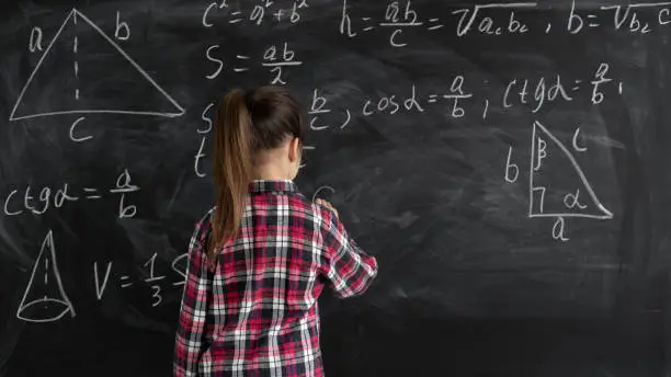 Photo of a schoolgirl girl in a shirt stands at the blackboard and writes mathematical formulas. concept back to school. Copy space.