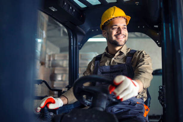 young caucasian male in working uniform and yellow hardhat operating forklift machine in warehouse storage room. - empilhadora imagens e fotografias de stock