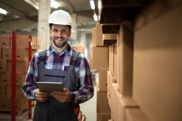 portrait of warehouse supervisor holding tablet computer and standing by cardboard boxes with goods in factory storage room. - warehouse worker imagens e fotografias de stock