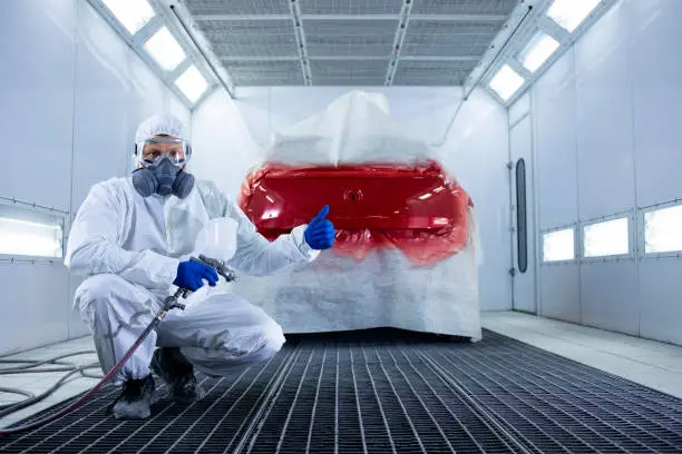 Photo of Portrait of professional car painter with protective clothing and mask holding thumbs up and standing by automobile in the painting chamber.