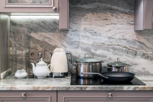 closeup of custom designed kitchen, with marble looking quartz countertop and backsplash. cream electric kettle with porcelain tea accessories on granite countertop next to ceramic hob with steel pots and pan primer plano de la cocina diseñada a medida, con encimera de cuarzo de aspecto de mármol y backsplash. hervidor eléctrico de crema con accesorios de té de porcelana en encimera de granito junto a vitrocerámica con ollas de acero y sartén
