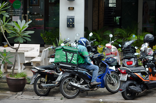 Thai delivery person is waiting on motorcycle in front of a condominium. In street many motorcycles are parked. Man is using mobile, scene is in Ladprao. Man is driving for Grab Food