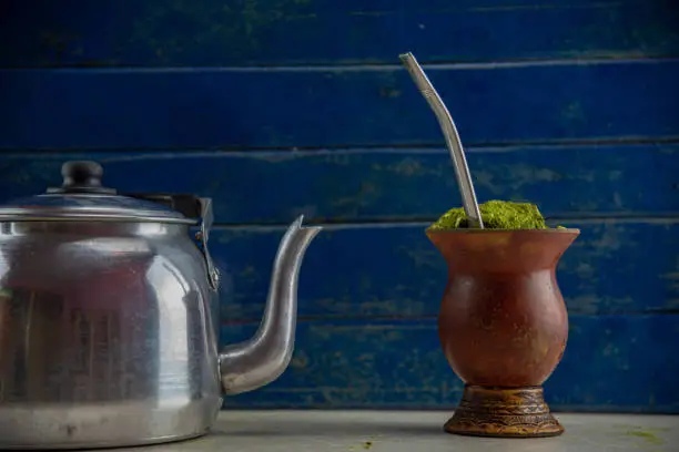 Photo of Chimarrão with Ilex paraguariensis herb and aluminum kettle on blue wooden background. Bitter mate