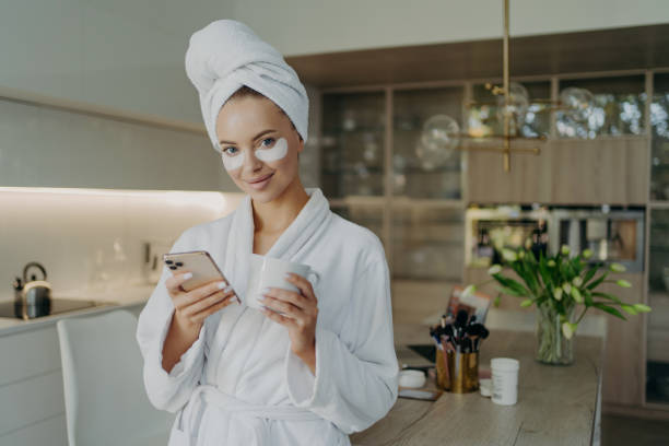 donna felice in accappatoio e asciugamano sulla testa rilassante con tazza di tè mentre fa procedure cosmetiche a casa - bathrobe women cup towel foto e immagini stock