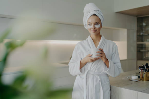 hermosa mujer en bata de baño y toalla en la cabeza bebiendo té mientras hace procedimientos de cuidado de la piel en casa - bathrobe women cup towel fotografías e imágenes de stock