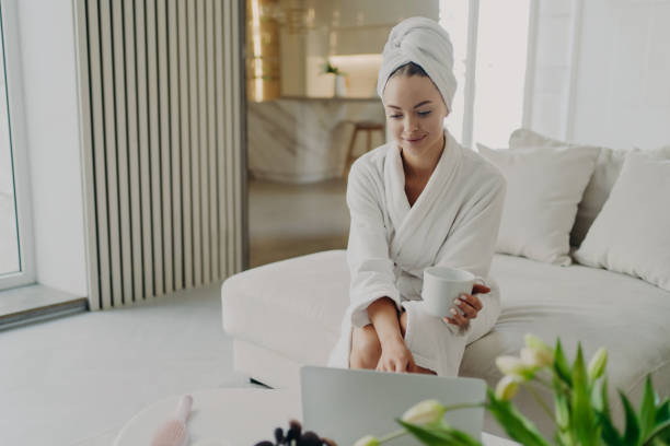 istock Young woman in white bathrobe working remotely on laptop while sitting on sofa in modern living room 1316481327