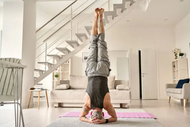 Strong fit sporty healthy old middle aged mature tattooed man doing headstand yoga morning fitness workout exercise standing on head on floor in living room at home. Seniors sport training concept.
