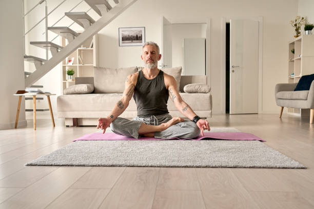 Calm healthy serene middle aged tattooed man meditating in yoga pose at home. Calm healthy serene old middle aged senior tattooed man meditating with eyes closed sitting in yoga pose in living room at home. Breathing exercises, meditation, no stress, peace of mind, zen concept. man yoga tattoo stock pictures, royalty-free photos & images