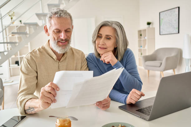 pareja de mediana edad leyendo facturas de papel calculando la pensión usando computadora portátil en casa. - parejas fotografías e imágenes de stock