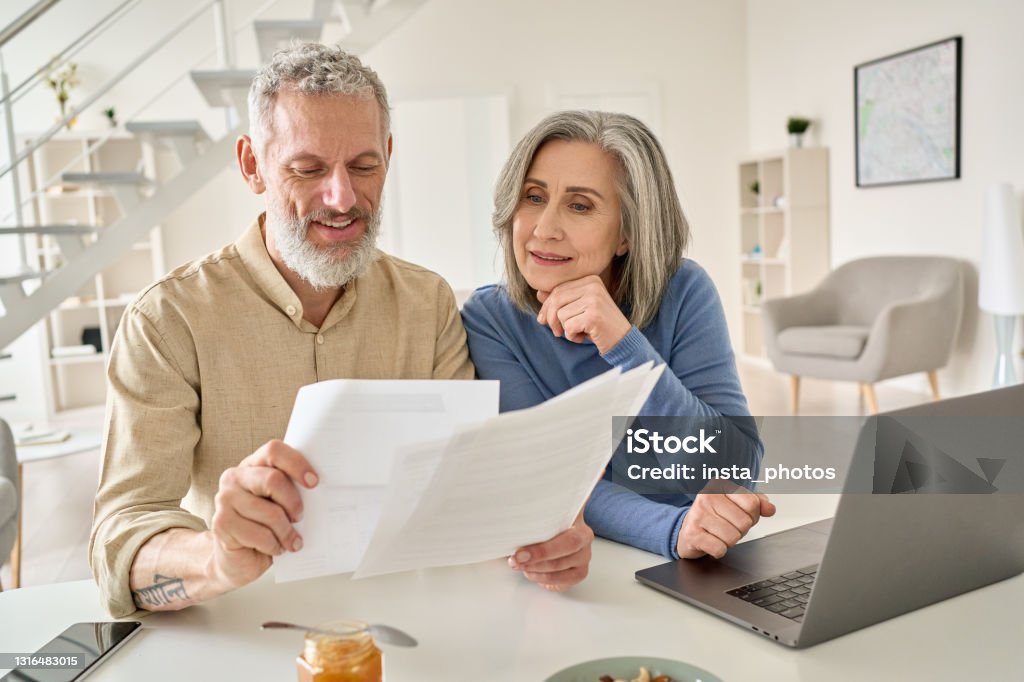 Para w średnim wieku czyta papierowe rachunki obliczające emeryturę za pomocą laptopa w domu. - Zbiór zdjęć royalty-free (Przejście na emeryturę)