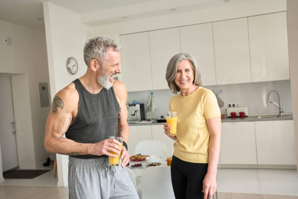 Healthy sporty happy seniors family couple drinking juice and laughing at home. Healthy sporty happy senior adults family couple drinking juice and laughing at home. Cheerful fit mid age older husband and wife enjoying talking, having fun standing in kitchen in the morning. 50 59 years stock pictures, royalty-free photos & images