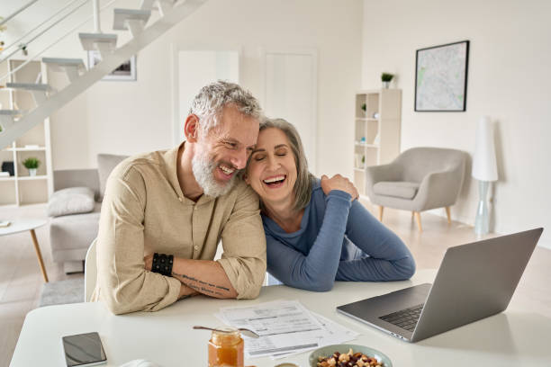 Happy mature older couple laughing, bonding sitting at home table with laptop. Happy mature older family couple laughing, bonding sitting at home table with laptop. Smiling middle aged senior 50s husband and wife having fun satisfied with buying insurance, paying bills online. 55 59 years stock pictures, royalty-free photos & images