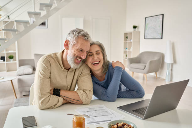 счастливая зрелая пожилая пара смеется, склеив сидя за домашним столом с ноутбуком. - retirement senior adult finance couple стоковые фото и изображения