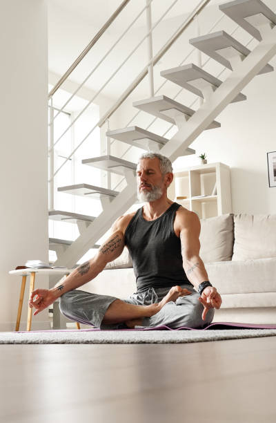 Healthy senior middle aged tattooed man meditating in yoga pose at home. Calm healthy serene old middle aged senior tattooed man meditating with eyes closed relaxing sitting in yoga pose in living room at home doing breathing exercises, feeling no stress, peace of mind. man yoga tattoo stock pictures, royalty-free photos & images