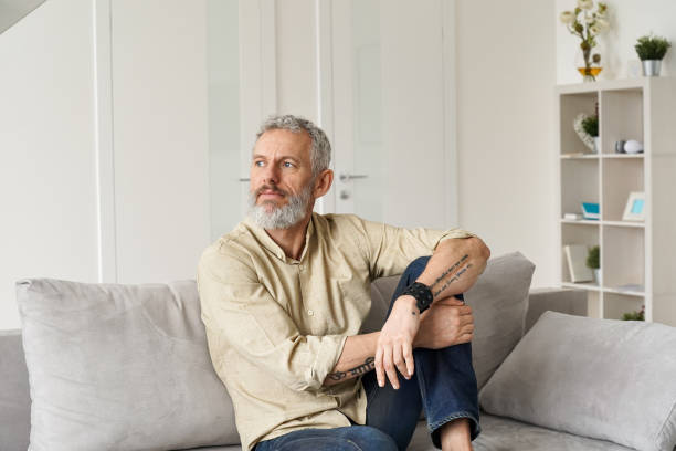 Relaxed mature thoughtful man relaxing sitting on sofa and thinking at home. Relaxed mature older thoughtful tattooed hipster man relaxing sitting on sofa and thinking at home. Pensive calm stylish mid aged single bearded guy resting on couch at home looking away, reflecting. introspection stock pictures, royalty-free photos & images