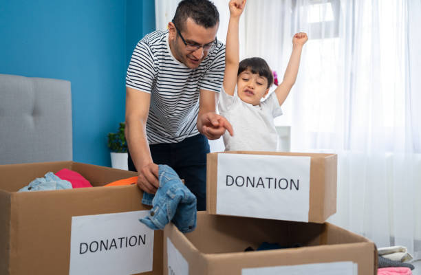 père et fils mignons mettant des vêtements et des jouets dans des boîtes pour des dons à la maison - charity and relief work donation box volunteer child photos et images de collection