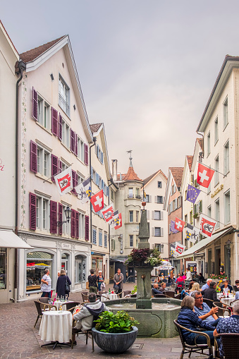 The Ochsenplatz, surrounded by rows of historic buildings, is one of the beautiful squares in Chur, the capital town of the Swiss canton of Graubunden. People are sitting at the outdoor tables of the cafés.