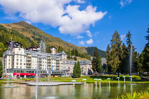Summer view of Davos, an Alpine resort town in the Swiss canton of Graubunden. People strolling in the public park with lake Seehofseeli, where stands the artwork 'Procession Sad' by Andreas Hofer.