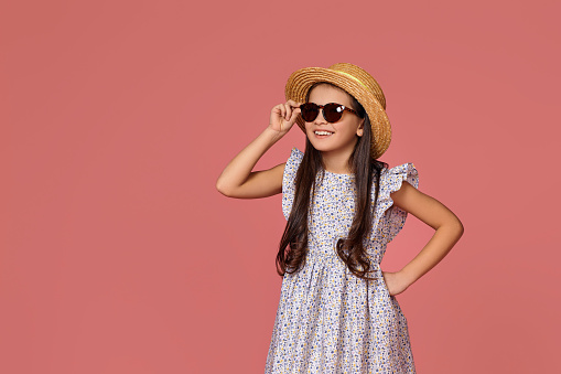 cute little child girl in summer dress, hat and sunglasses looking at the side on pink background with copy space. Holiday concept