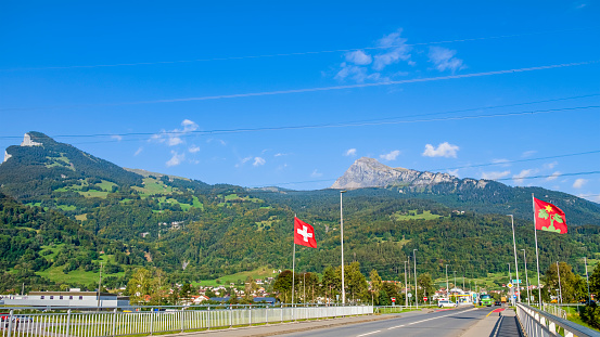 The flags of Switzerland and the municipality of Wartau wave on the bridge crossing the Rhine river.