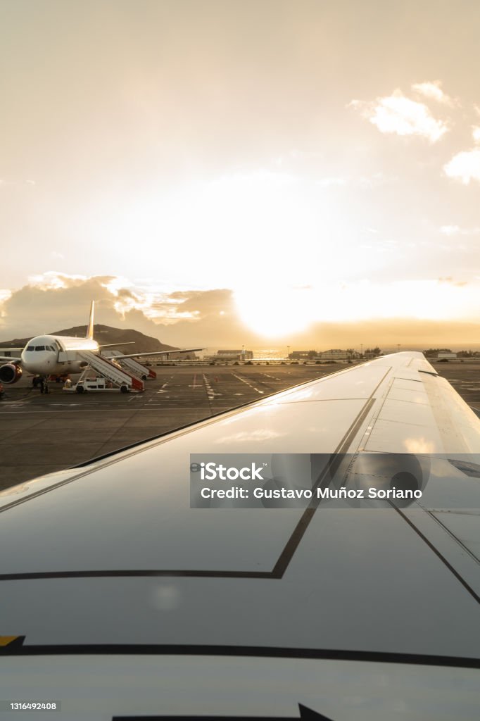 Wing of a plane in the foreground on the runway with a plane in the background ready to board at sunset Wing of a plane in the foreground on the runway with a plane in the background ready to board at sunset. Travel concept Aerospace Industry Stock Photo