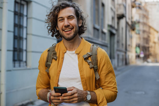 happy young man on the street, smiling and holding smart phone - young men men one young man only casual imagens e fotografias de stock