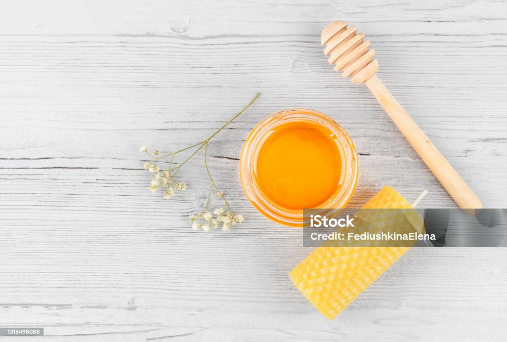 Beekeeping products on a white wooden background. Beekeeping products. Open jar with honey, beeswax candle, wooden  honey dipper and a flower on a white wooden background. Top view, copy space. Alternative medicine, vegetarianism, healthy eating. Beeswax Stock Photo