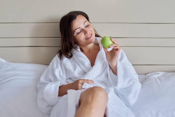 istock Middle-aged woman in white bathrobe with green apple in bed 1316499044