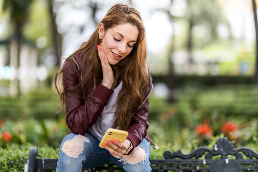 Young girl writing a message with her smartphone sitting on a bench in the park format with copy space close up