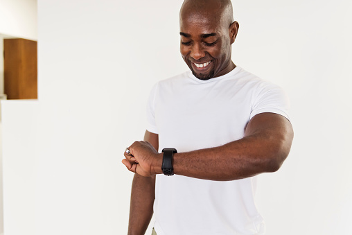 Mature adult man looking at his training watch at home in small apartment. He is from African descent, wearing sport clothing with white top. White background. Horizontal waist up indoors shot with copy space.