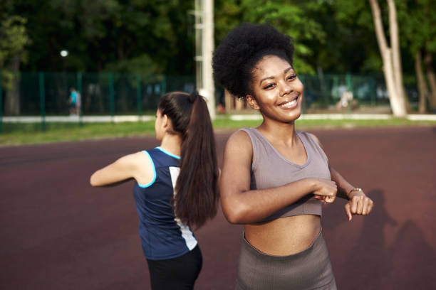 две подтянутые улыбающиеся молодые женщины, тренируясь вместе в парке, выполняя упражнения на растяжку и разгибание рук, чтобы улучшить ги� - physical activity стоковые фото и изображения