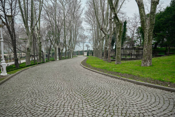Cobblestone way and path in Gulhane public park with white street lamps and withered and dried trees and green grass during overcast sky and rain and bosporus background.