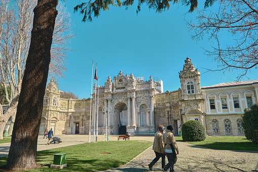 istanbul, Turkey. Old ottoman patterned and baroque style dolmabahce palace during sunny day and facade of entrance gate made of marble stone with walking tourists and visitors with mask.