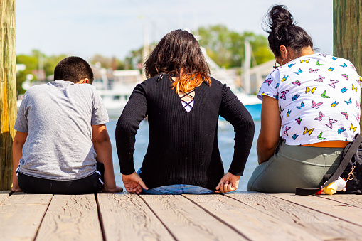 Annapolis, MD, USA 05-02-2021: A hispanic family with mother in the middle and her son and daughter on both sides are sitting on the city dock of Annapolis looking at the sea. They are enjoying the beautiful sunny weather.