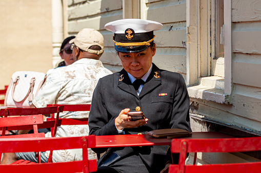 Annapolis, MD, USA 05-02-2021: A young Asian American female cadet training at the US Naval Academy in Annapolis is sitting at a restaurant table with full uniform. She is looking at her phone.