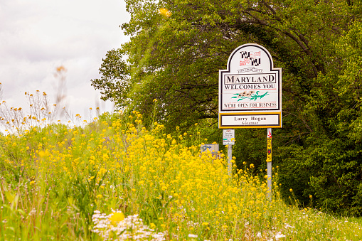 Point of Rocks, MD, USA 04-30-2021: Maryland Welcomes You road sign on the scenic byway US Route 15 at the border of Maryland and Virginia.  It has MD flag and says open for business. Wildflowers in spring are all over the place