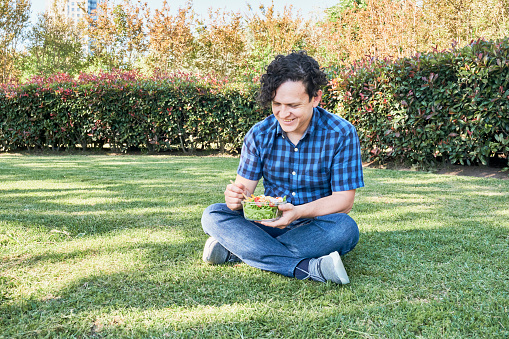 Smiling young man sitting on the grass eating a fresh salad. Healthy living and eating concepts.