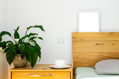 A decorated with blooming peace lily in a ceramic flowerpot and a tea or coffee cup on a saucer wooden bedside table by wooden bed. head of the bed is decorated with a white frame with copy space.