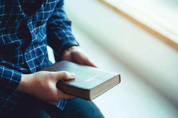 mann sitzt beim auskunden von bibel oder buch über betonwand mit fensterlicht - bible holding reading book stock-fotos und bilder