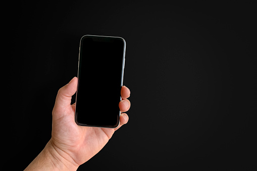 Man holding mobile phone with blank screen on black background, closeup of hand. Space for text