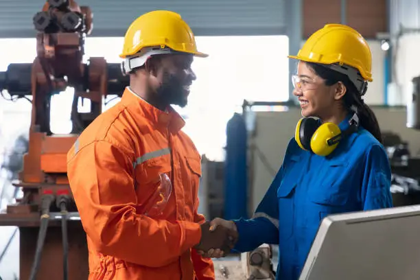 African male engineer and asian female engineer standing smiling eye contact and shakehands together in side of robotic arm in factory.