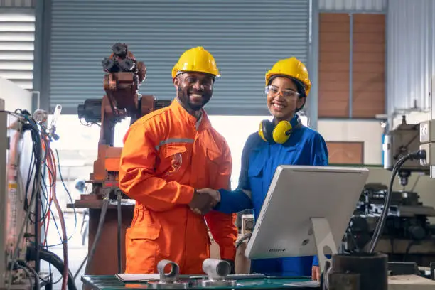 African male engineer and asian female engineer looking at camera smiling  and shakehands together in side of robotic arm in factory.