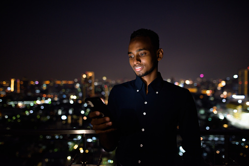 Portrait of handsome African businessman outdoors at rooftop in Bangkok, Thailand using mobile phone