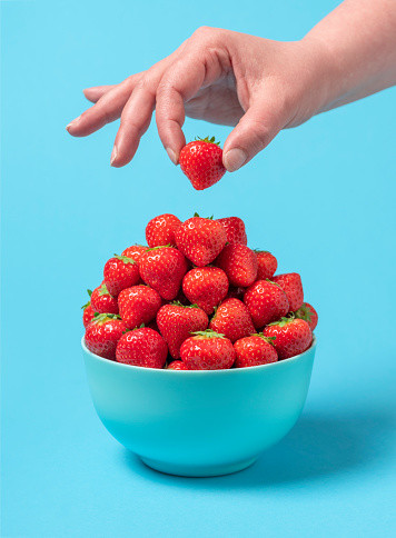 Bowl with fresh organic strawberries isolated on a blue background. Woman's hand taking a strawberry.