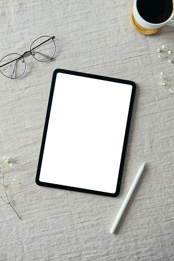 Blank tablet screen mockup, drawing pencil, glasses, cup of coffee on linen fabric. Elegant feminine workspace top view.